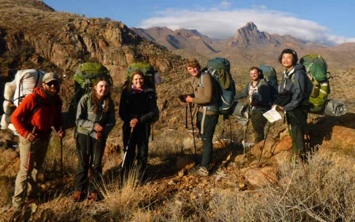 a group of students carrying backpacks stand amidst a desert landscape with mountains in the background